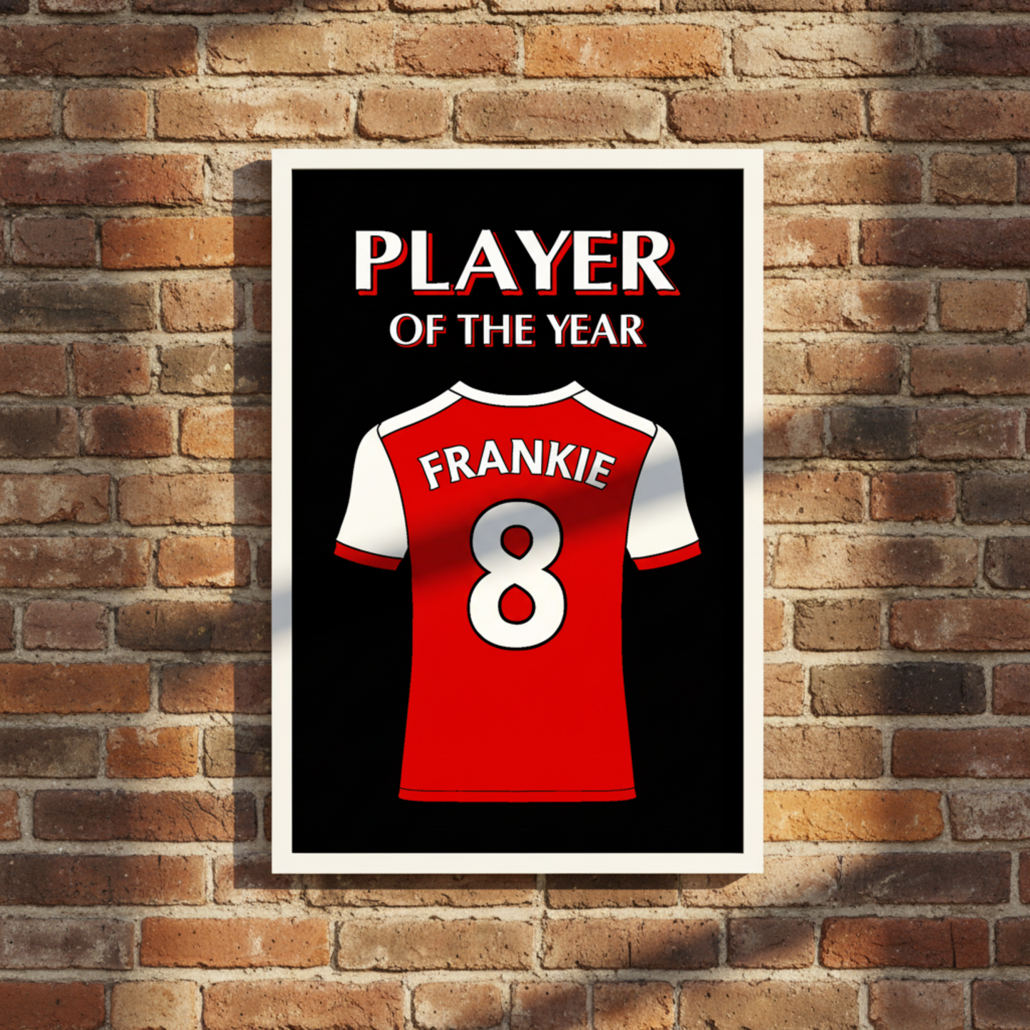 A personalised football shirt featuring a red and white football jersey with the name 'FRANKIE' and number '8' on the back, set against a black background with the text 'PLAYER OF THE YEAR' in white and red. The print is framed and displayed on a rustic brick wall.