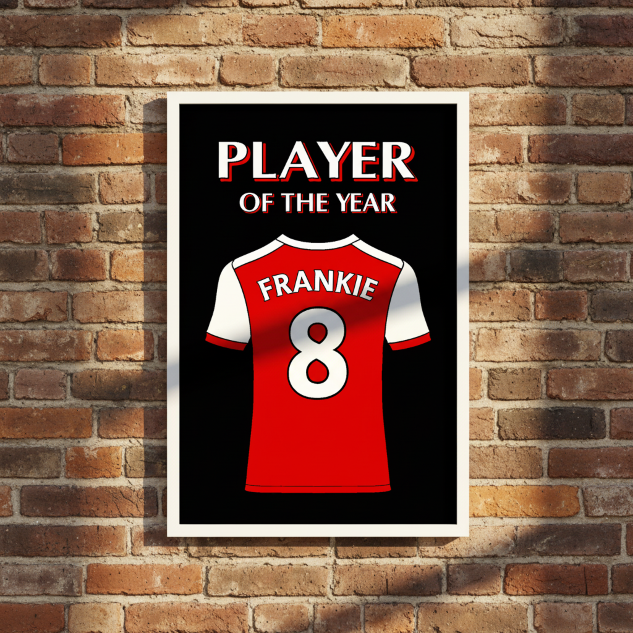 A personalised football shirt featuring a red and white football jersey with the name 'FRANKIE' and number '8' on the back, set against a black background with the text 'PLAYER OF THE YEAR' in white and red. The print is framed and displayed on a rustic brick wall.