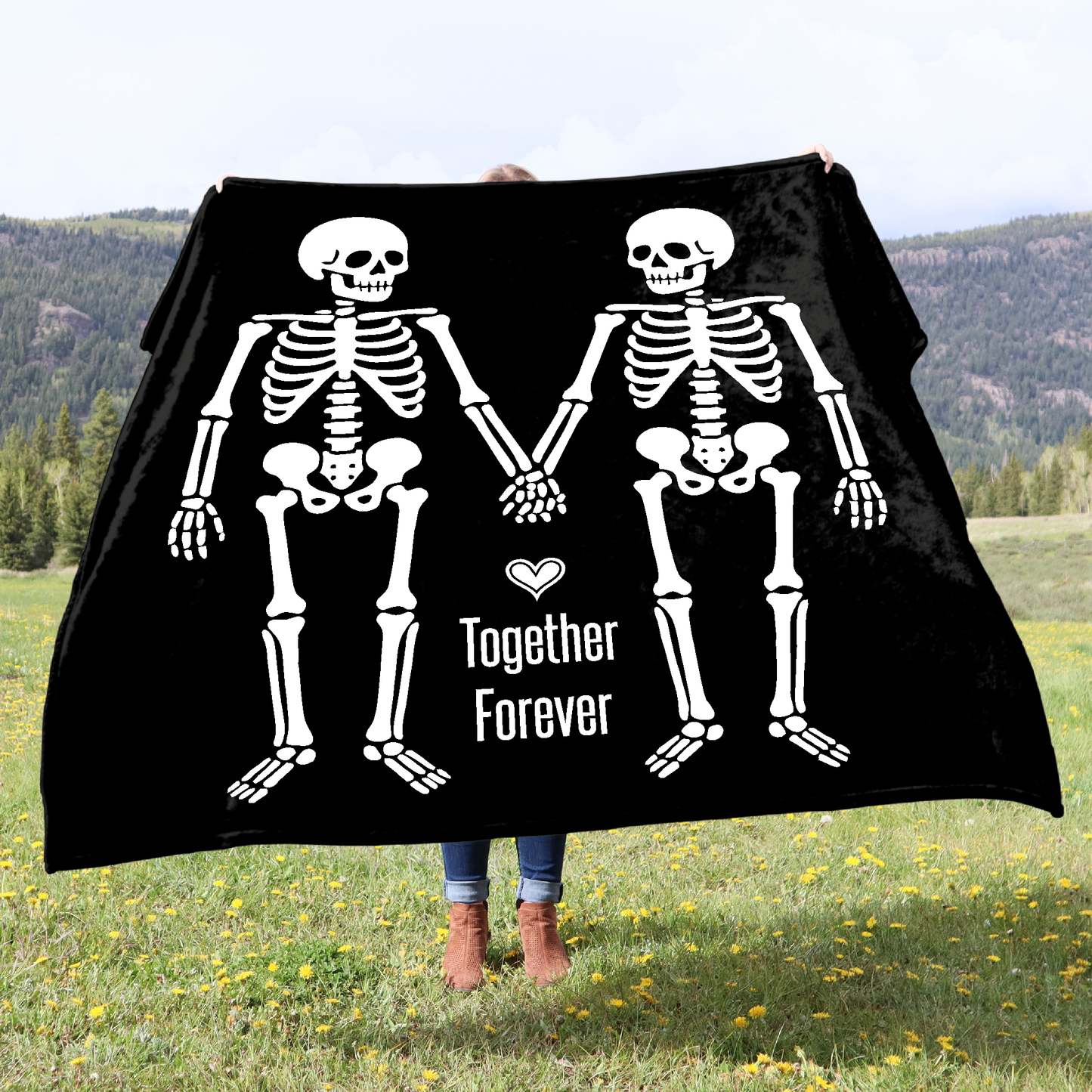 Person holding a black blanket with skeleton design and 'Together Forever' text in a scenic outdoor setting.